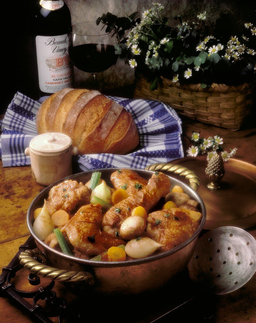 Coq au Vin in einem Kupfertopf, dahinter Brot und Rotwein