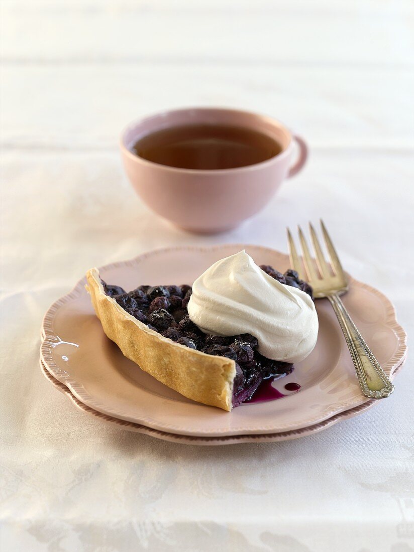 Piece of blueberry pie with cream in front of cup of tea