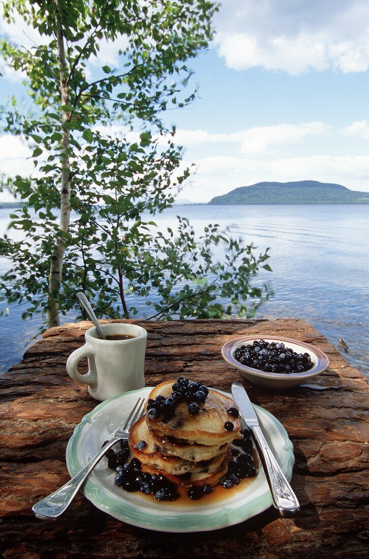 Pancakes mit Heidelbeeren und Kaffeetasse auf Mauer am See