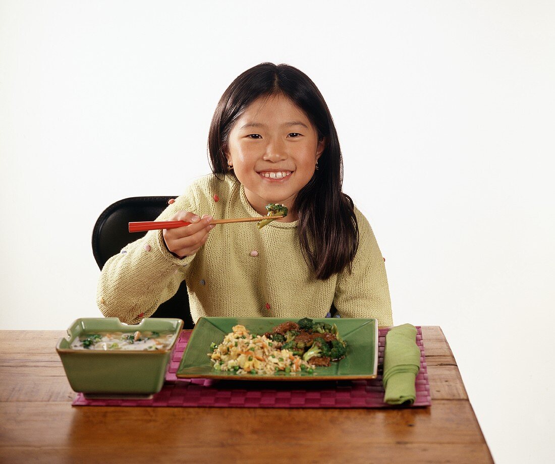 Child Sitting at Table Eating Broccoli Stir Fry and Fried Rice With Chopsticks, Bowl of Soup