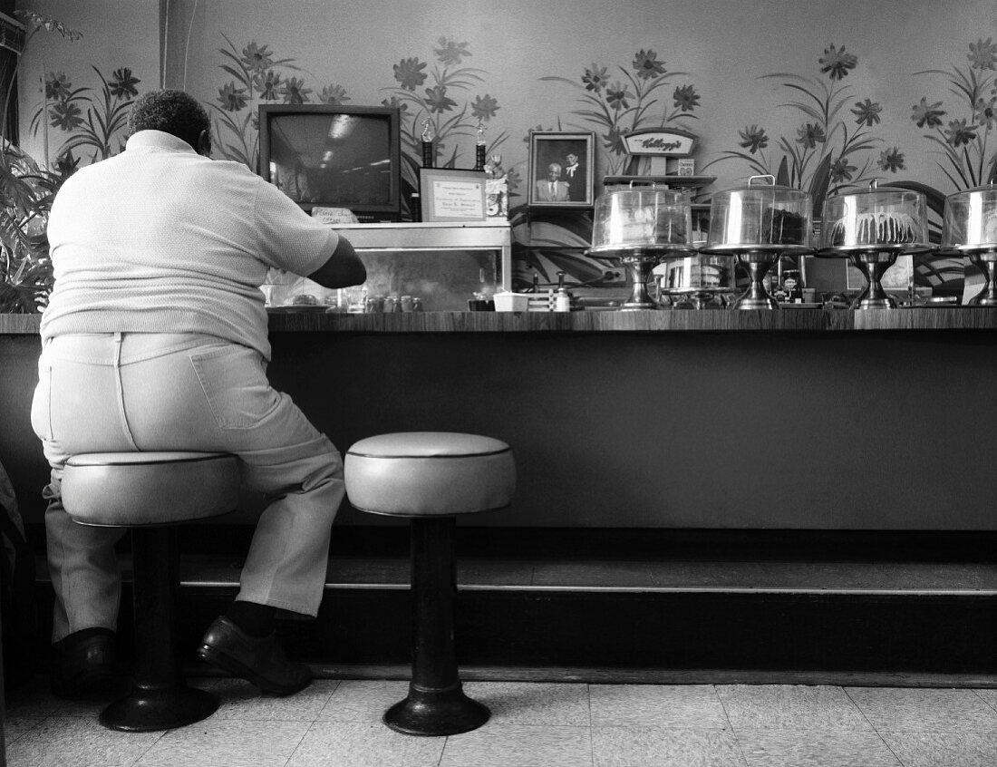 A Large Man at a Lunch Counter with a Variety of Cakes