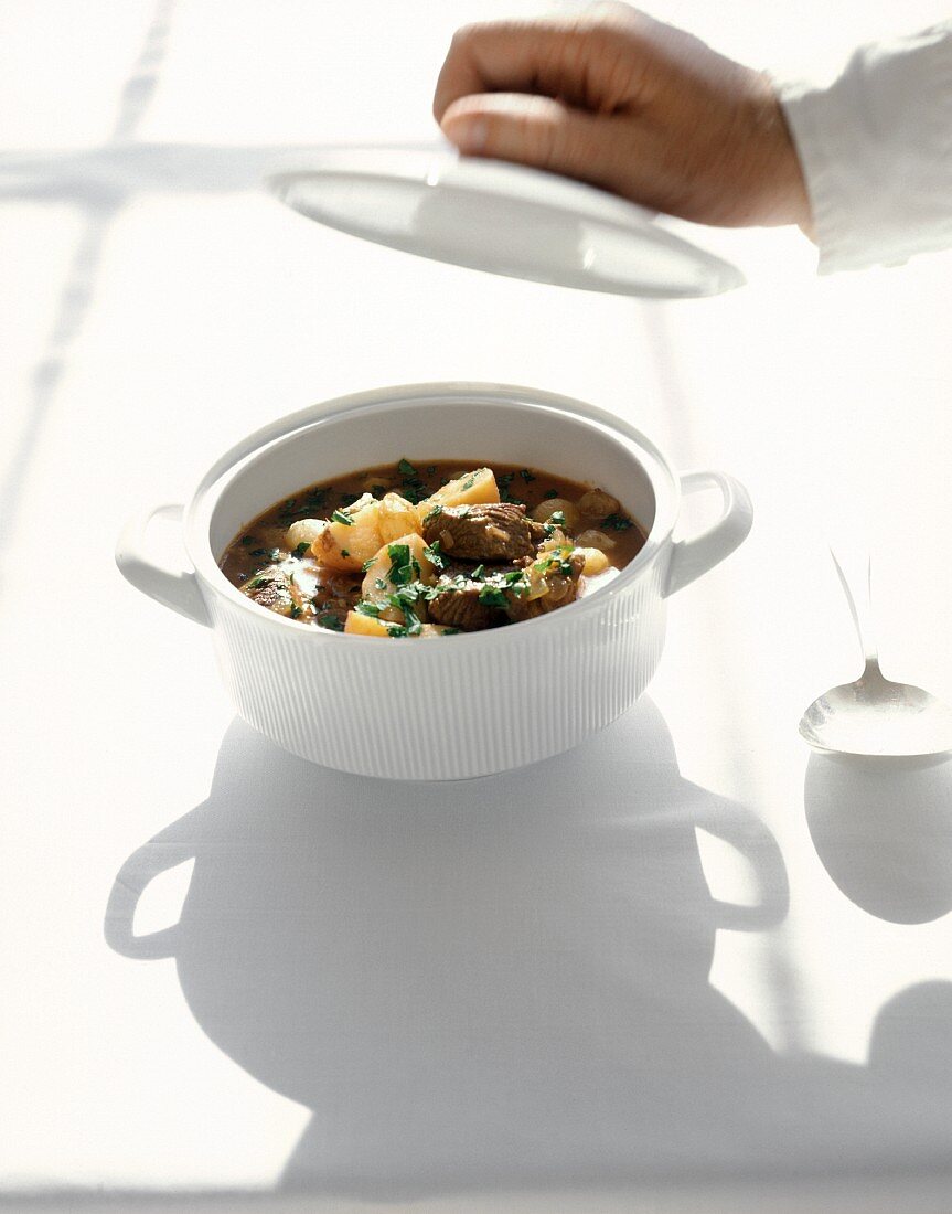 Meat stew in soup tureen, hand holding lid above it