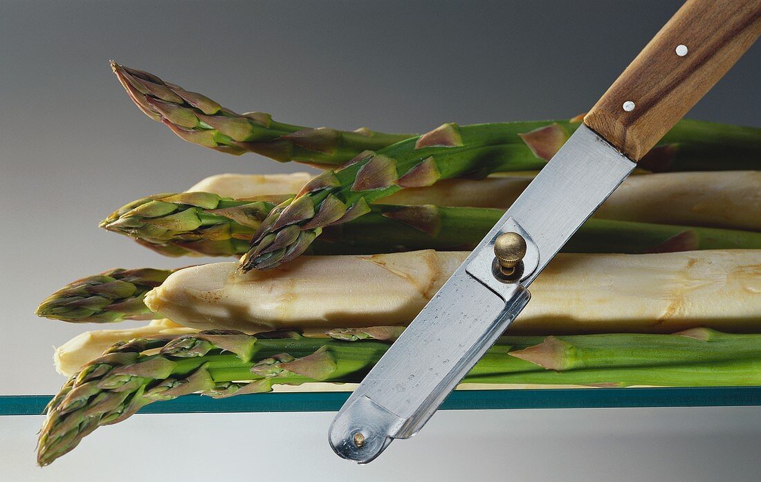 Green and white asparagus spears on sheet of glass, peeler