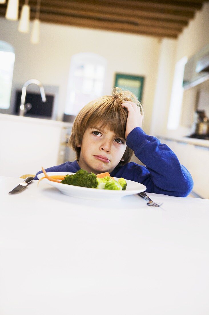 Kleiner Junge sitzt unglücklich vor Gemüseteller in der Küche