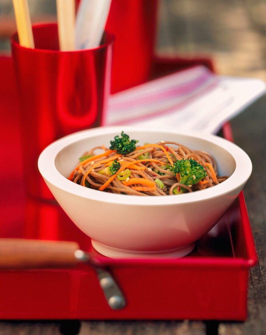 A Bowl of Soba Noodles with Broccoli, Carrots and Scallions on a Red Tray