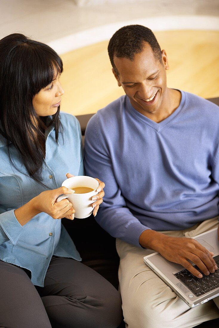Woman with cup of coffee beside man with computer on sofa