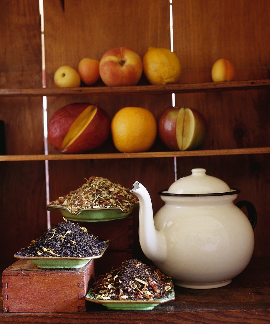 A Variety of Tea Leaves next to a Tea Pot with Fruit Behind