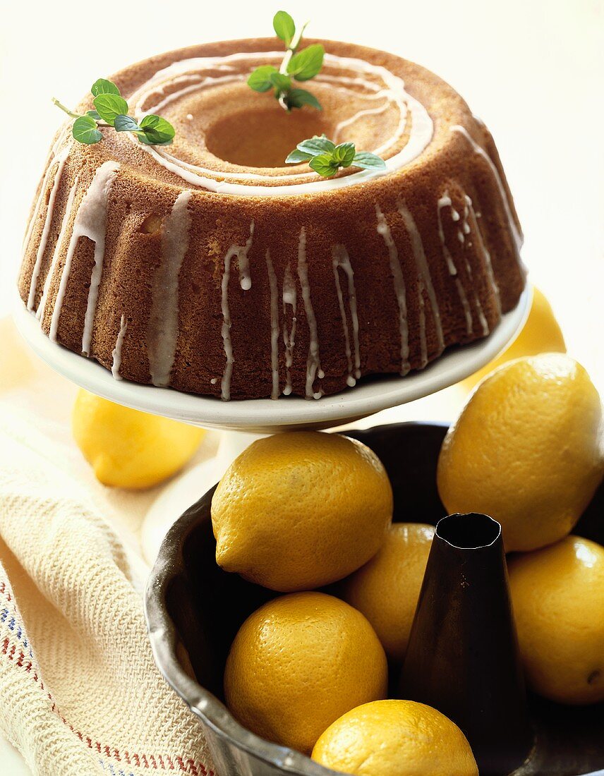 Glazed Lemon Bundt Cake on Cake Pedestal with Fresh Lemons in Baking Pan
