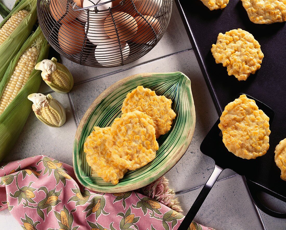 Corn Fritters on a Plate; Spatula Removing Corn Fritters From Skillet; Fresh Corn and Eggs