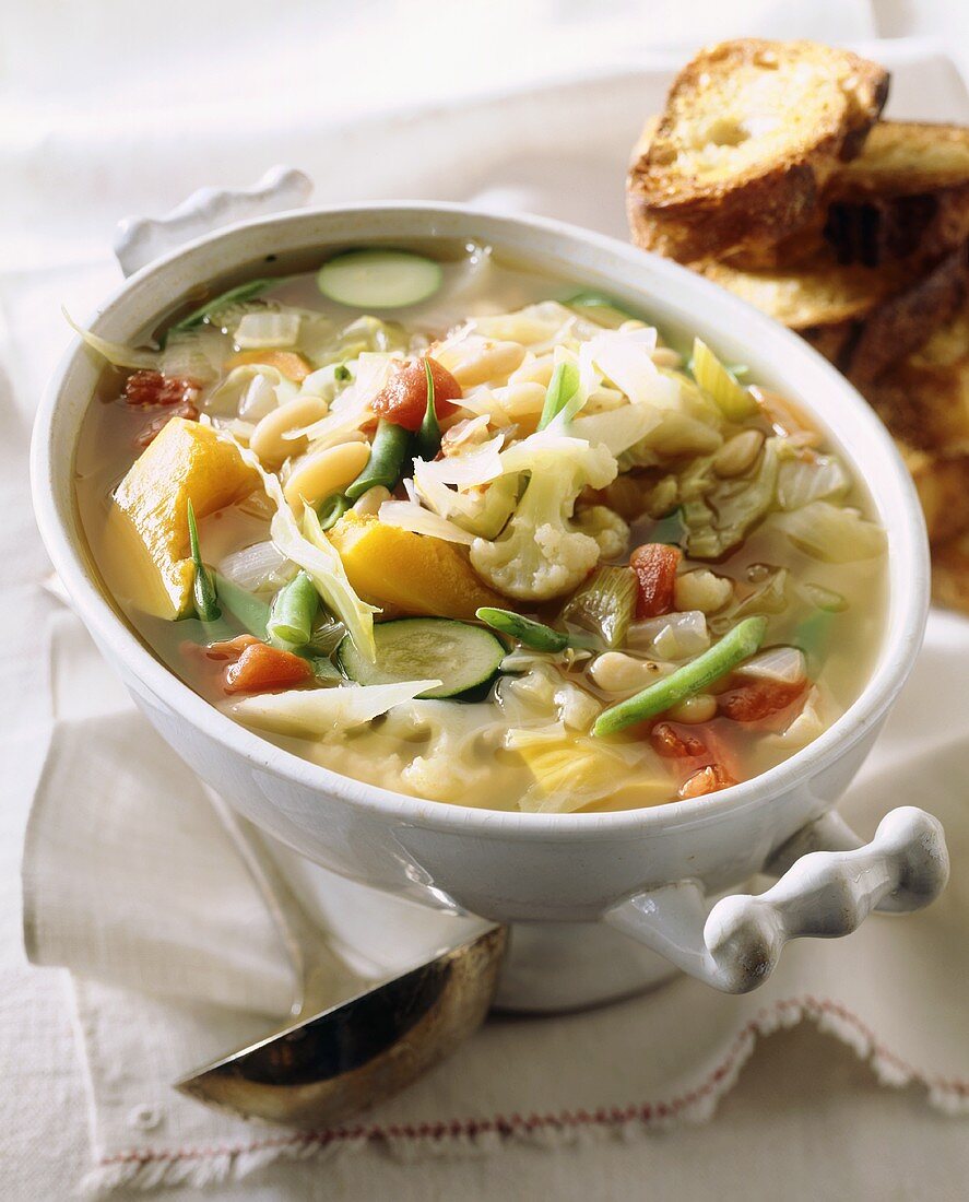 Gemüsesuppe in Suppenterrine, daneben Kelle und Brot