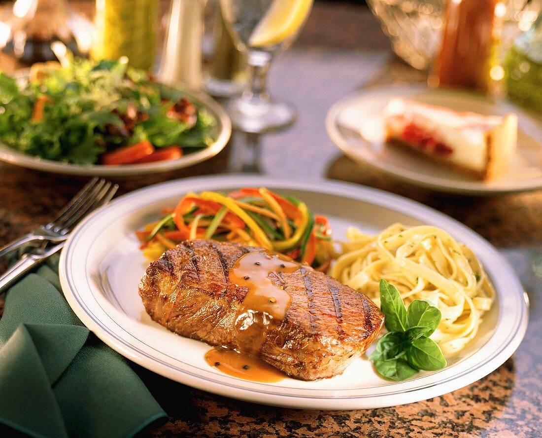 Grilled New York Steak with Fettucini on Table with Side Salad and Cheesecake