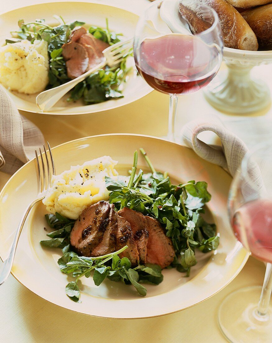 Plates of Sliced Lamb Over Watercress with Mashed Potatoes; Glasses of Red Wine