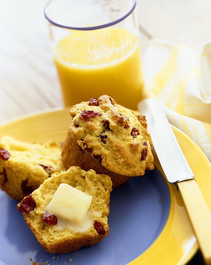 Cranberry Corn Muffins on a Plate; One Sliced in Half with Butter Melting on It; Glass of Orange Juice