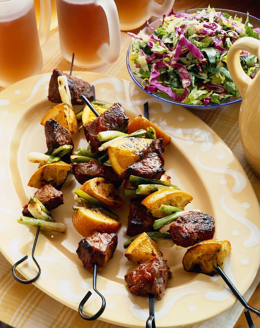 Beef, Orange and Green Onion Kabobs on a Platter; Side Salad and Mugs of Beer