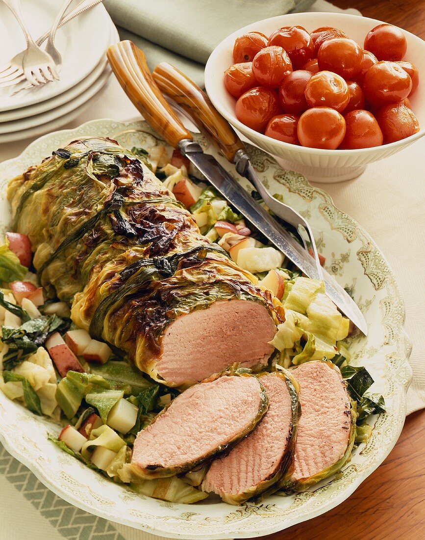 Partially Sliced Cabbage Wrapped Pork Roast on a Platter with Cabbage and Potatoes; Bowl of Tomatoes