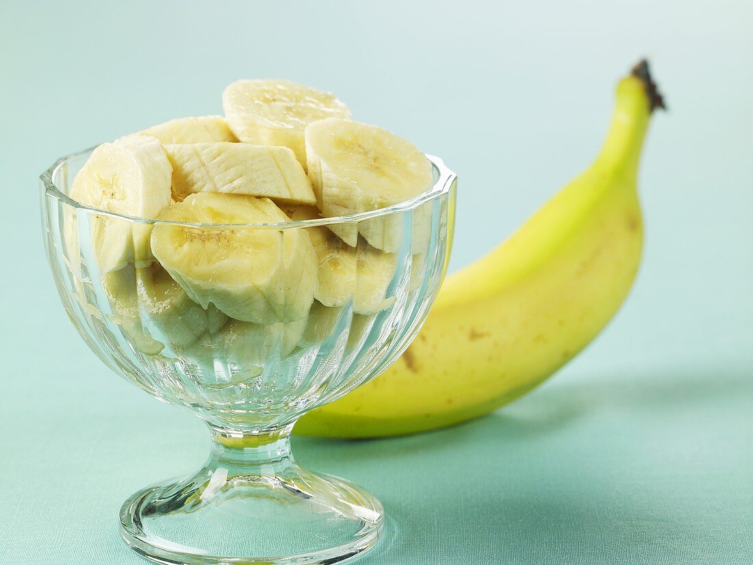 Bananenscheiben in Glasschale, im Hintergrund ganze Banane