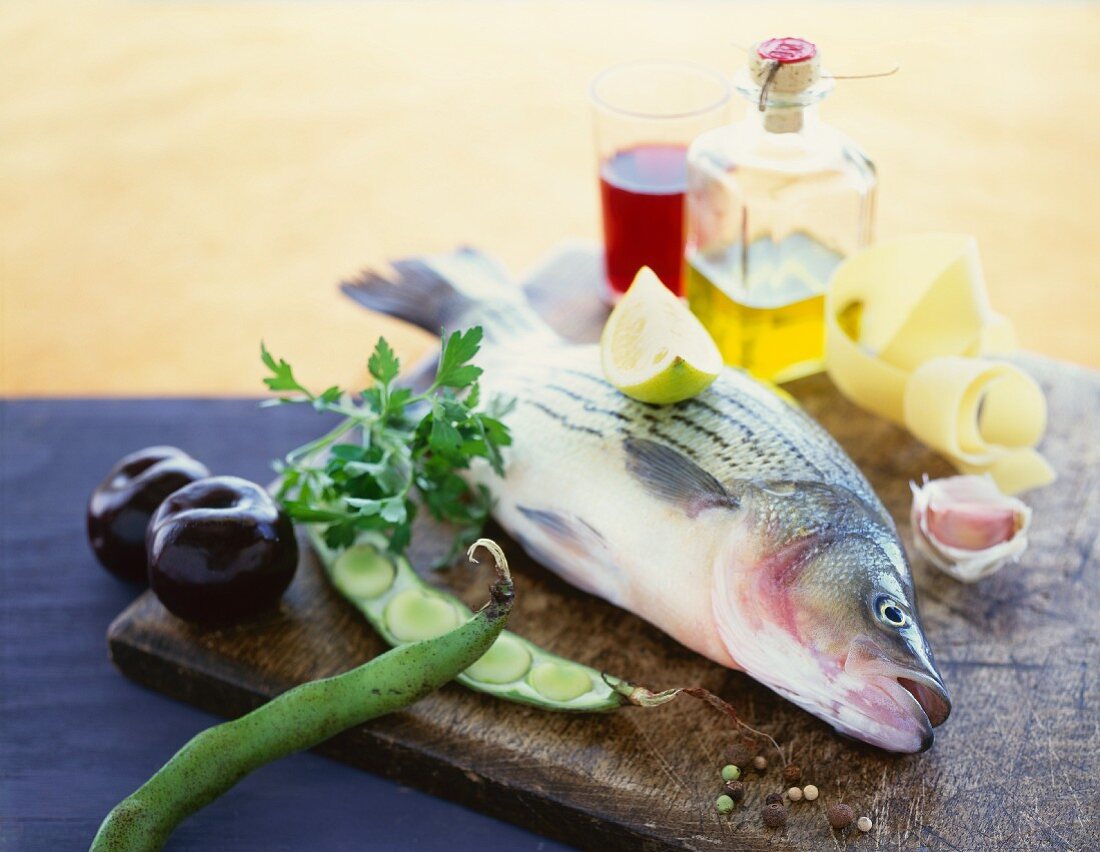 Assorted Fresh Ingredients on a Cutting Board; Whole Fish, Plums, Beans, Garlic, Olive Oil and Vinegar