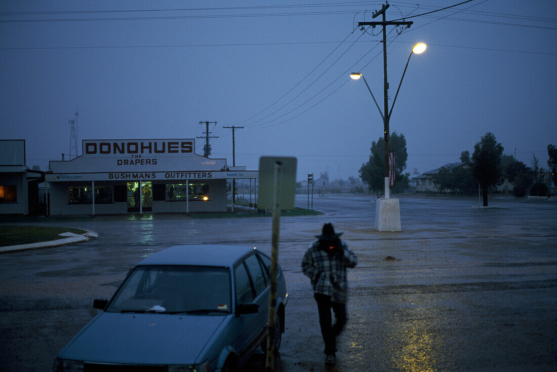 Mann geht nachts im Regen über einen verlassenen Parkplatz, Boulia, Queensland, Australien