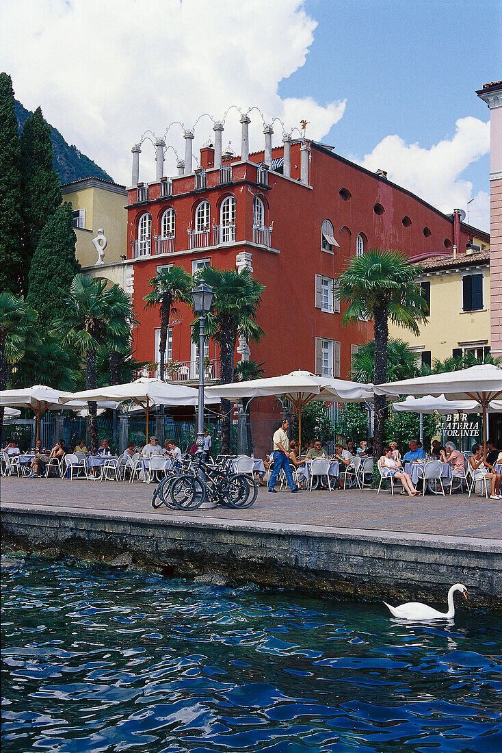 Menschen sitzen unter Sonnenschirmen vor einem Café an der Promenade, Torbole, Gardasee, Trentino, Italien, Europa