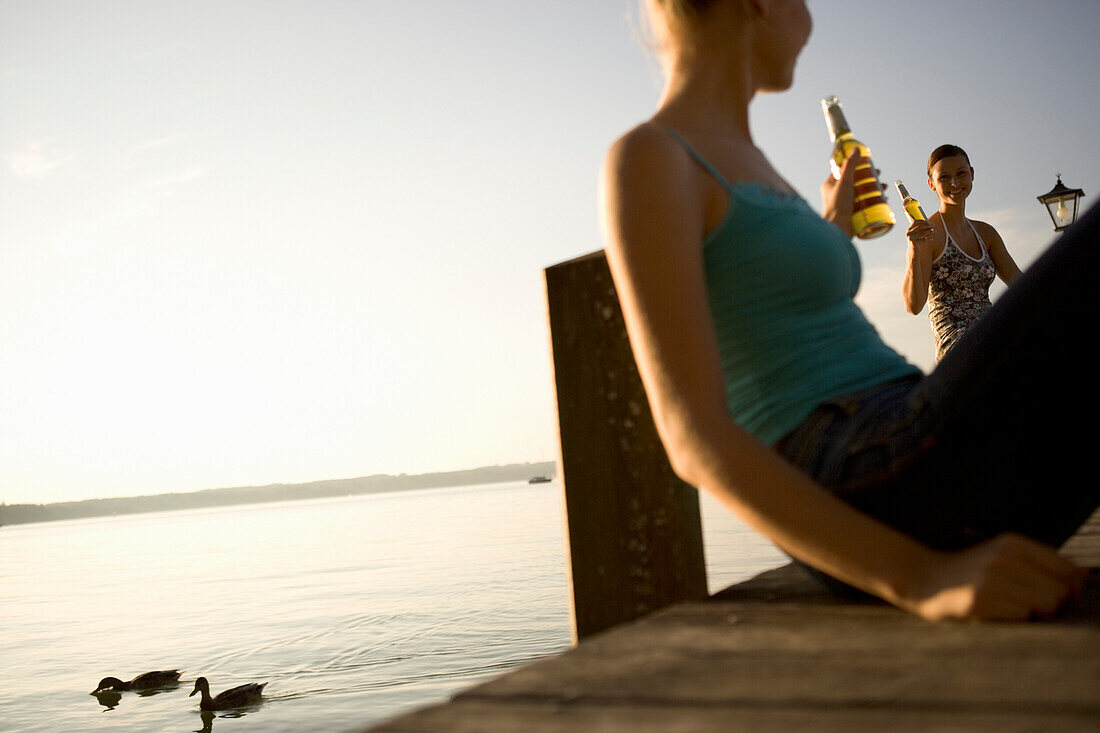 Young women sitting on the jetty with a bottle of beer, Lake Starnberg, Bavaria, Germany