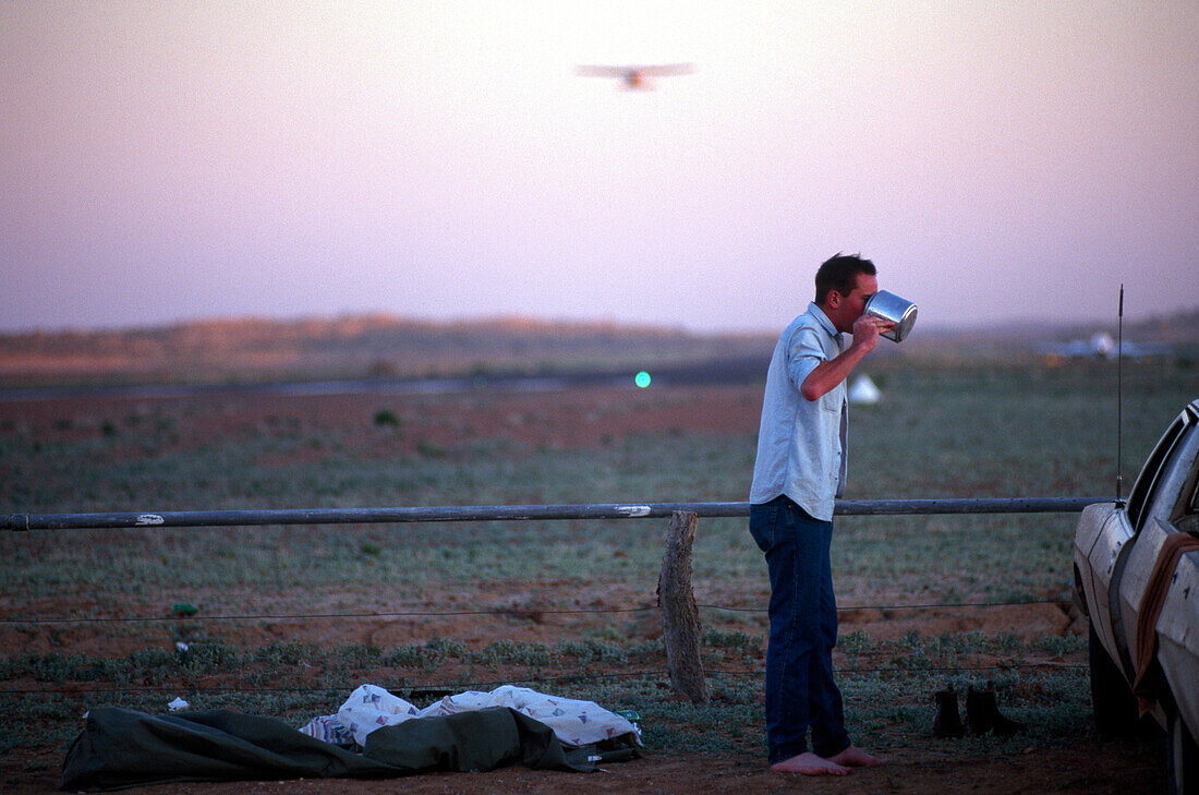 Junger Mann trinkt morgens Tee vor der Startbahn des Birdsville Airport, Birdsville, Simpson Desert, Australien