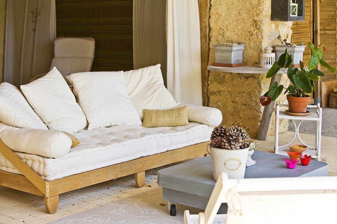Sofa mit einfachem Holzgestell und großen, weissen Kissen auf der Terrasse eines rustikalen Landhauses