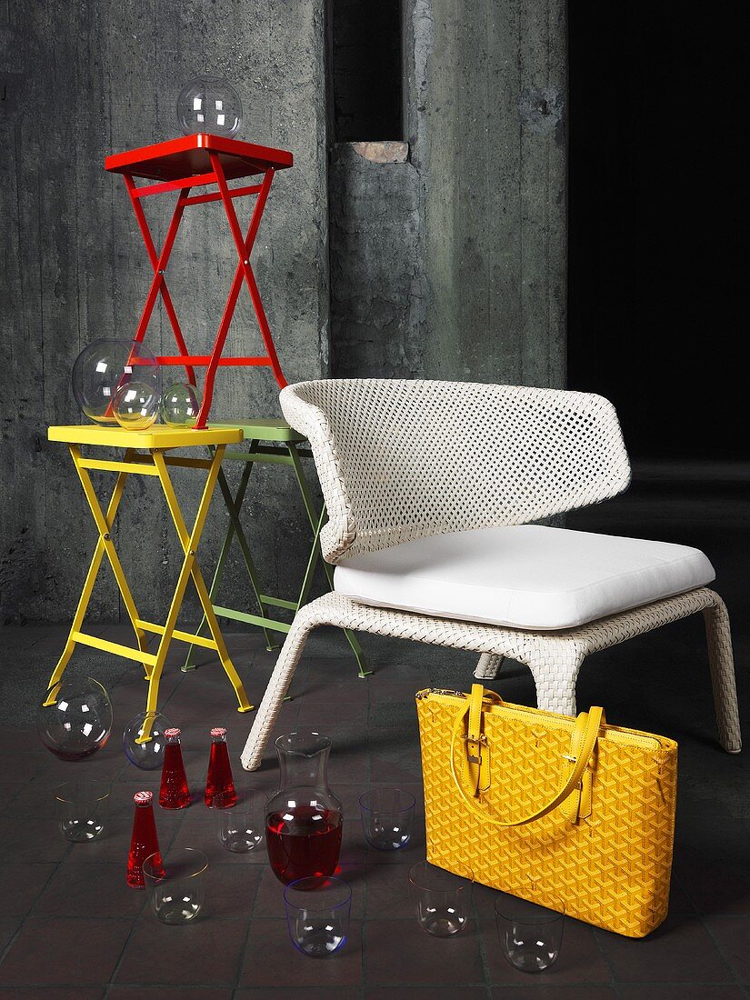 Glasses and vases arranged on and around a white wicker chair and a stack of coloured tables