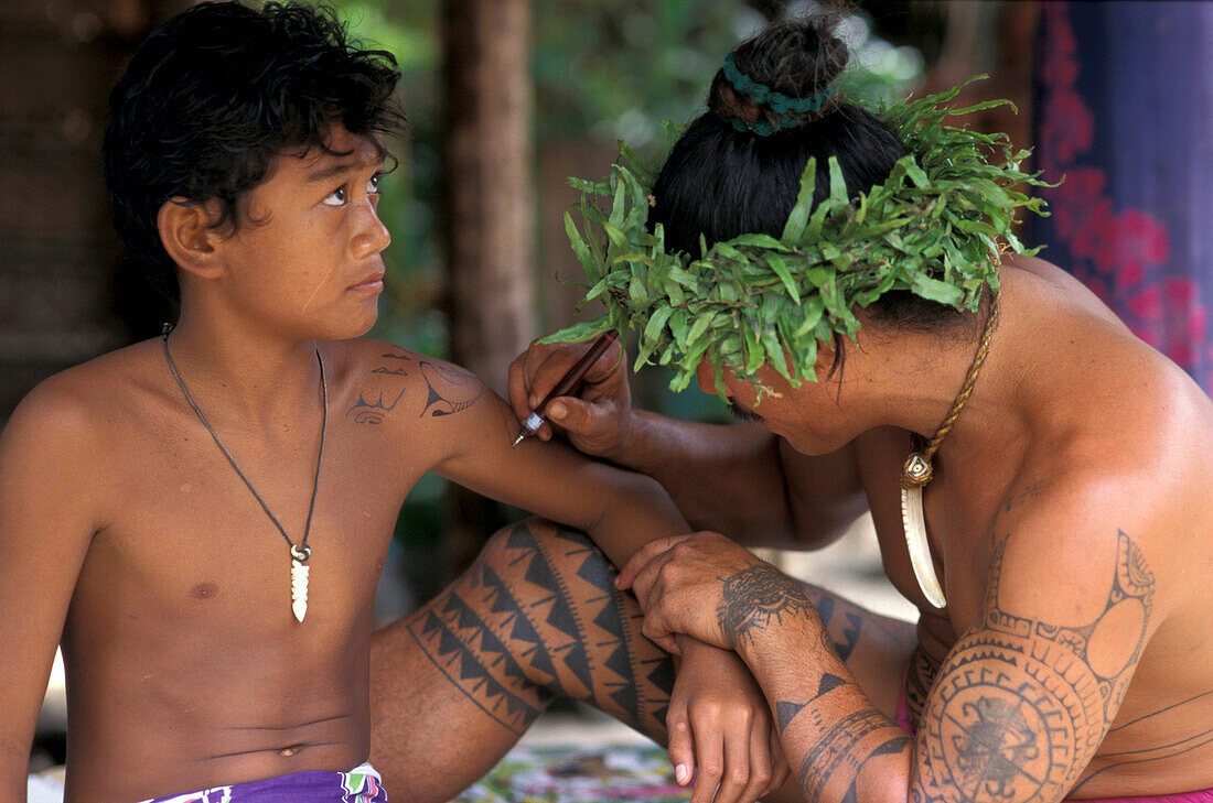 Ein Mann mit Kopfschmuck tätowiert einen Jungen im Dorf Tiki, Moorea, Französisch Polynesien, Ozeanien