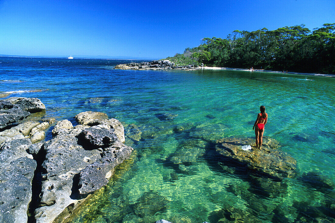 Eine Frau steht auf einem Felsen in einer Bucht unter blauem Himmel, Honeymoon Bay, Jervis Bay Marine Park, New South Wales, Australien