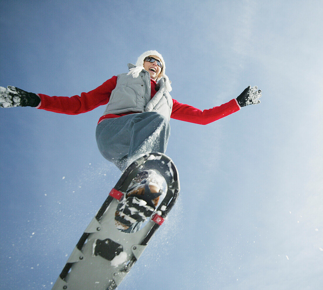 Junge Frau springt mit Schneeschuhen im Winter gegen blauen Himmel und roten Pullover und weißer Wollmütze