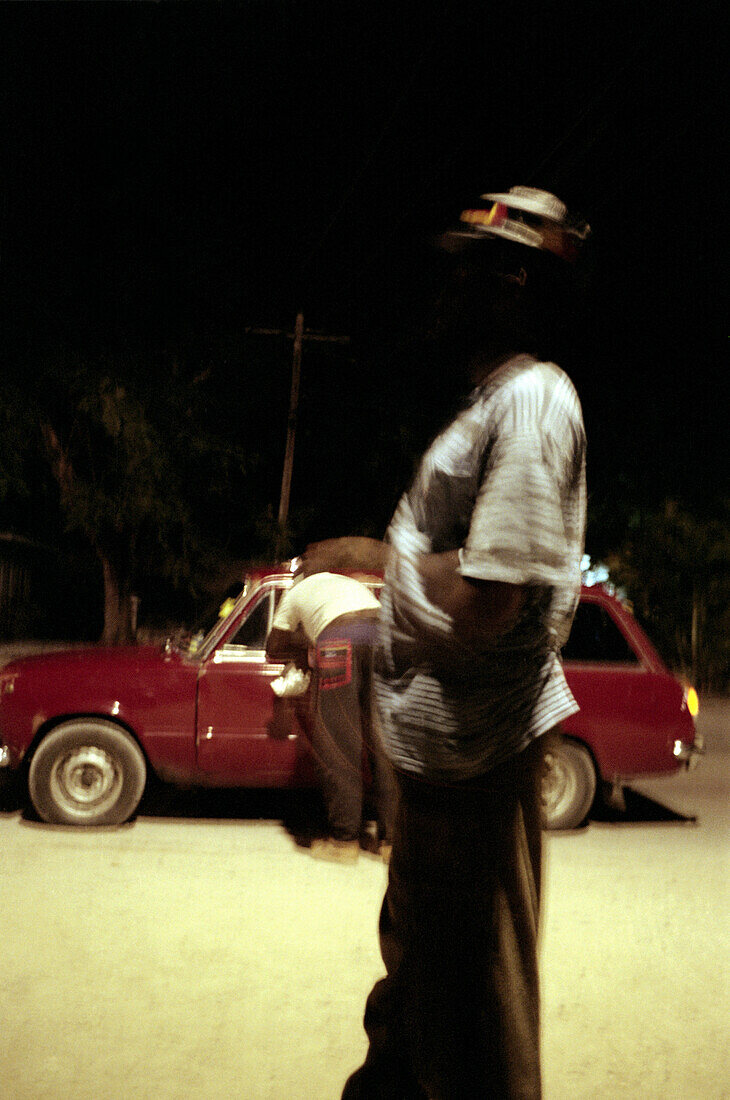 Two jamaican men standing in front of a red car at night, Jamaica, Carribean