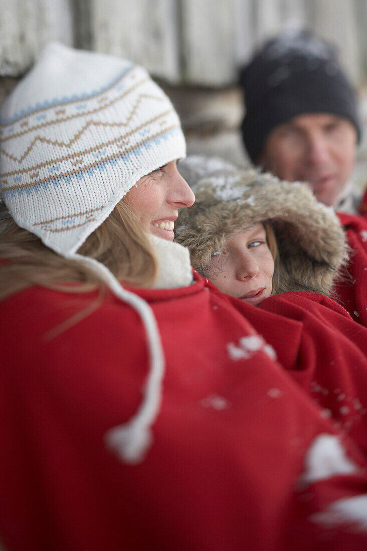 Parents and son leaning on wooden hut, warming with red blanket