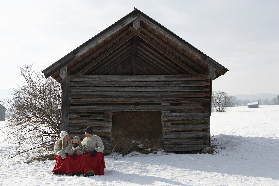 Parents and son sitting near wooden hut, warming with red blanket