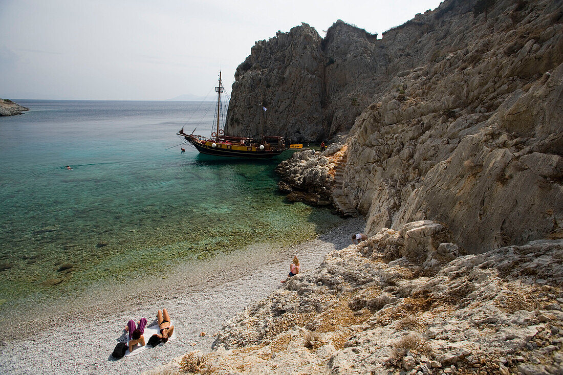People lying at beach of a bay at Kalymnos next to a sailing boat, Greece