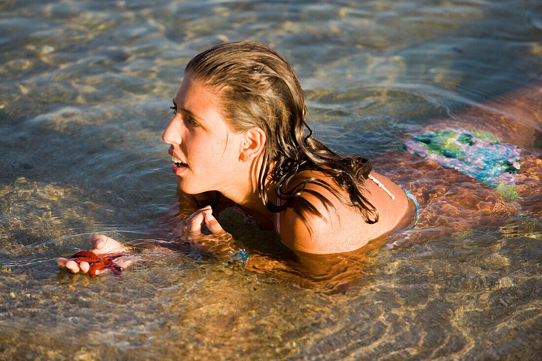 Woman lying in shallow water at Paranga Beach and holding a starfish in her hand, Mykonos, Greece