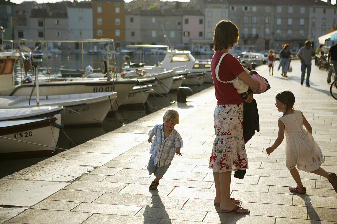 Girl and boy running round their mother, Cres Harbour, Cres Island, Croatia