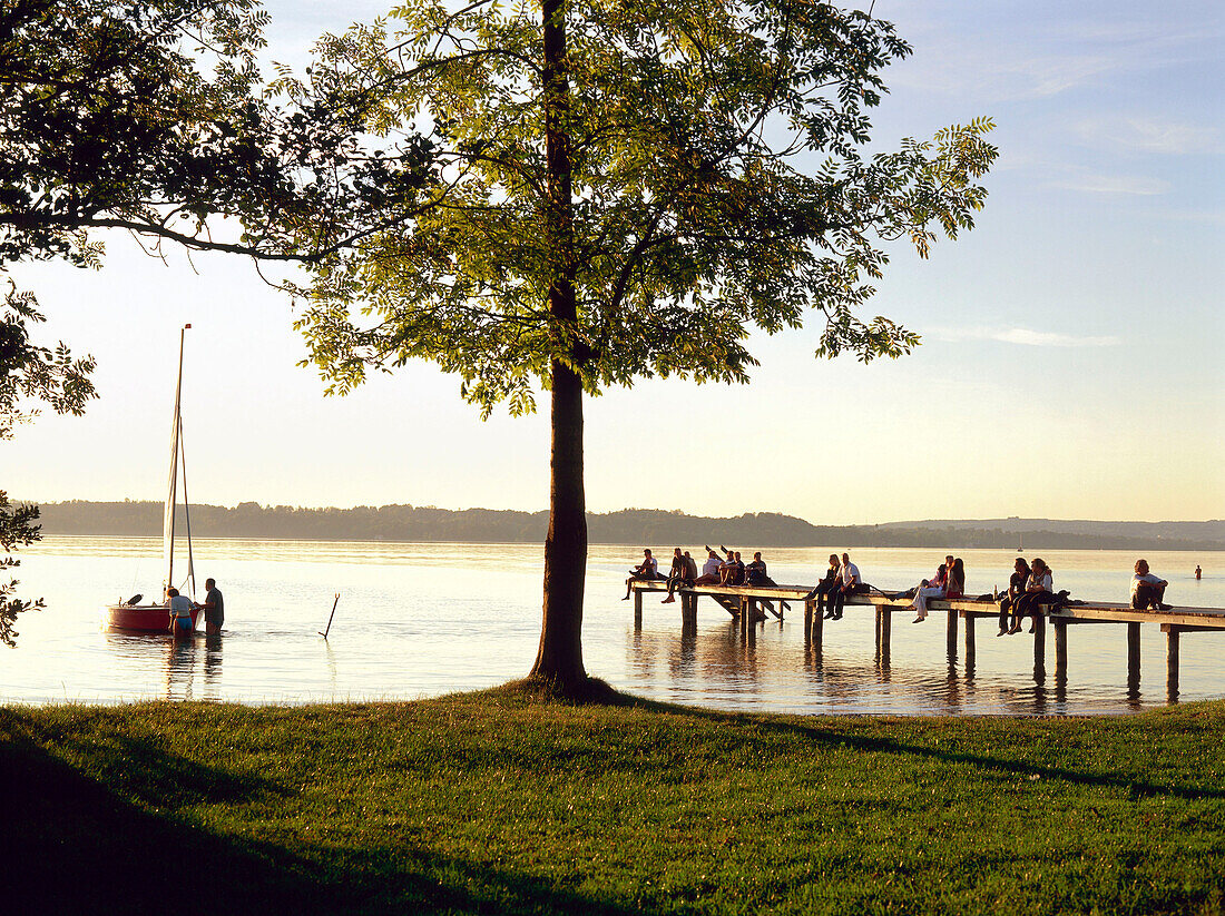 Menschen sitzen auf Holzsteg, St. Heinrich, Starnberger See, Oberbayern, Deutschland