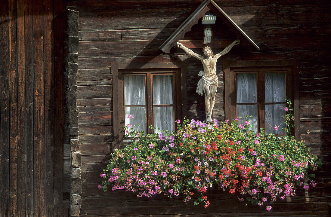 Kruzifix mit Blumenschmuck an Hauswand eines Blockhauses aus Holz, Defreggental, Kärnten, Österreich