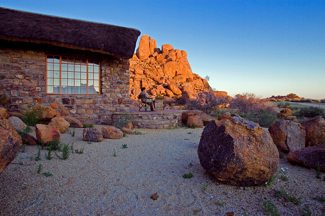 Junger man sitzt vor einem Bungalow und betrachtet die von der Abendsonne beschienenen Felsen. Canon Lodge, Gondwana Canon Park. Fish River Canyon, Südliches Namibia, Afrika. MR.