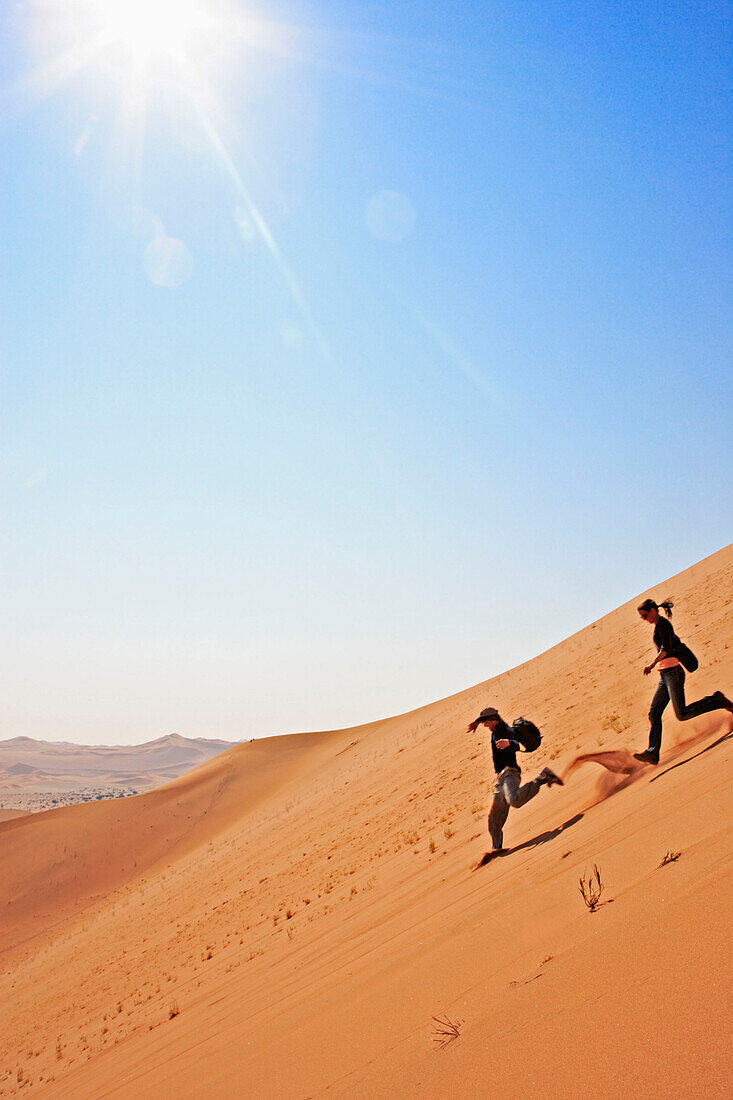 Eine junge Frau und ein junger Mann rennen über die steile Flanke des Big Daddy hinunter. Eine der höchsten Dünen der Erde. Die Sossusvlei Dünen. Namib Wüste. Namibia. Afrika. MR