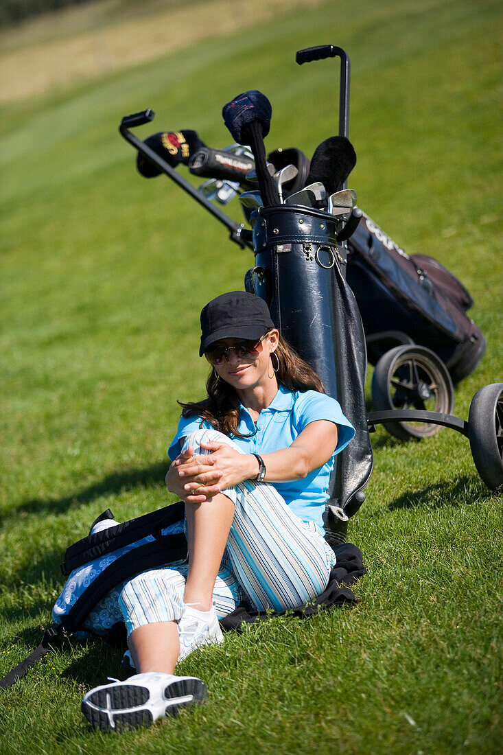 Junge Frau sitzt im Gras lehnt an Golftasche