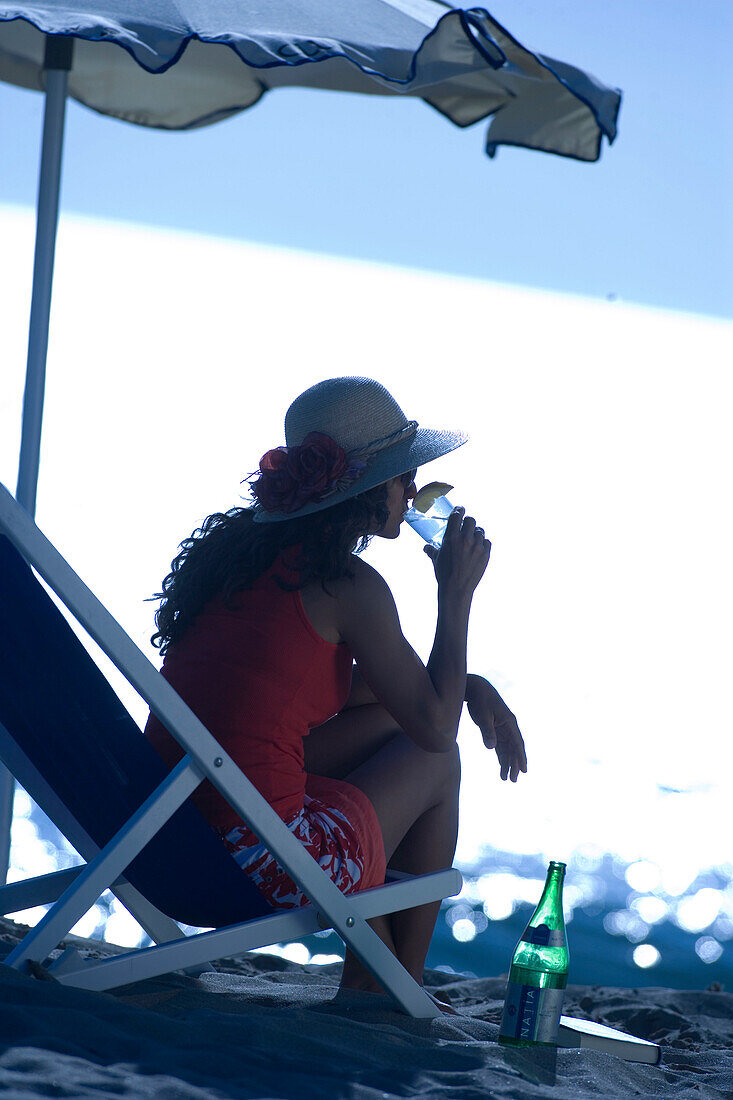 Woman sitting in deck chair on beach and drinking, Apulia, Italy