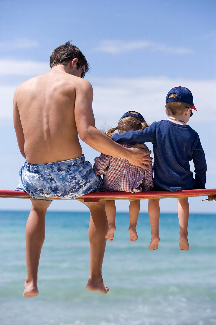 Junger Mann und zwei Kinder sitzen auf einer Bank am Strand, Apulien, Italien