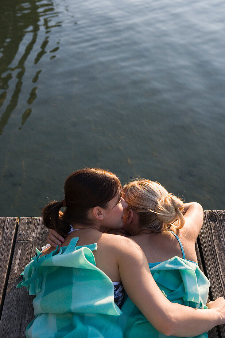 Two young women lying on jetty in blanket, whispering