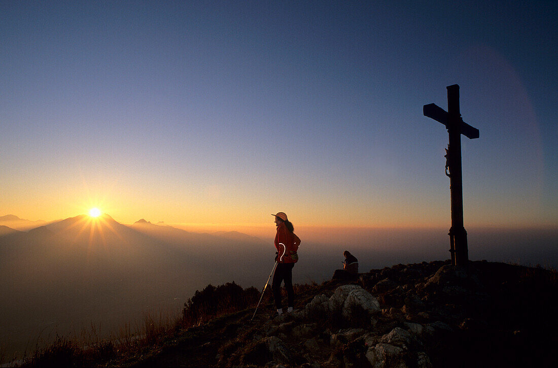 Bergsteigerin am Gipfelkreuz des Heubergs mit Sonnenuntergang über der Wendelsteingruppe, Bayerische Voralpen, Oberbayern, Bayern, Deutschland