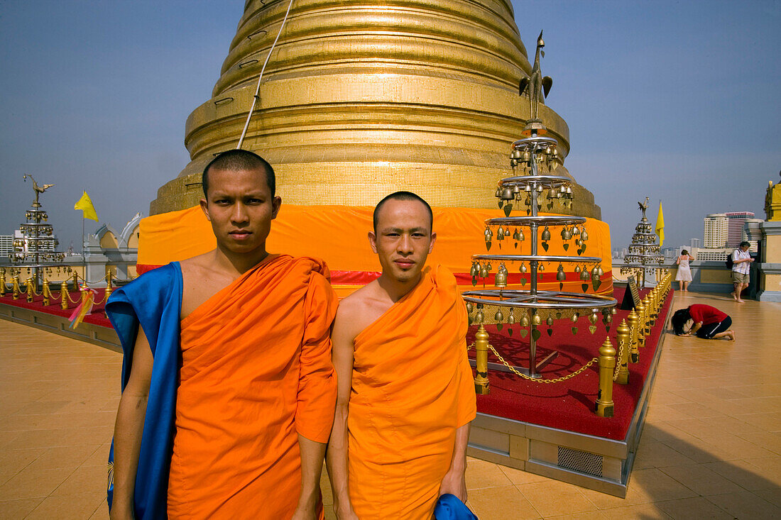 Two monks in front of gilded Chedi, housed a Buddha relic of the Wat Saket on the Golden Mount, woman praying in background, Bangkok, Thailand