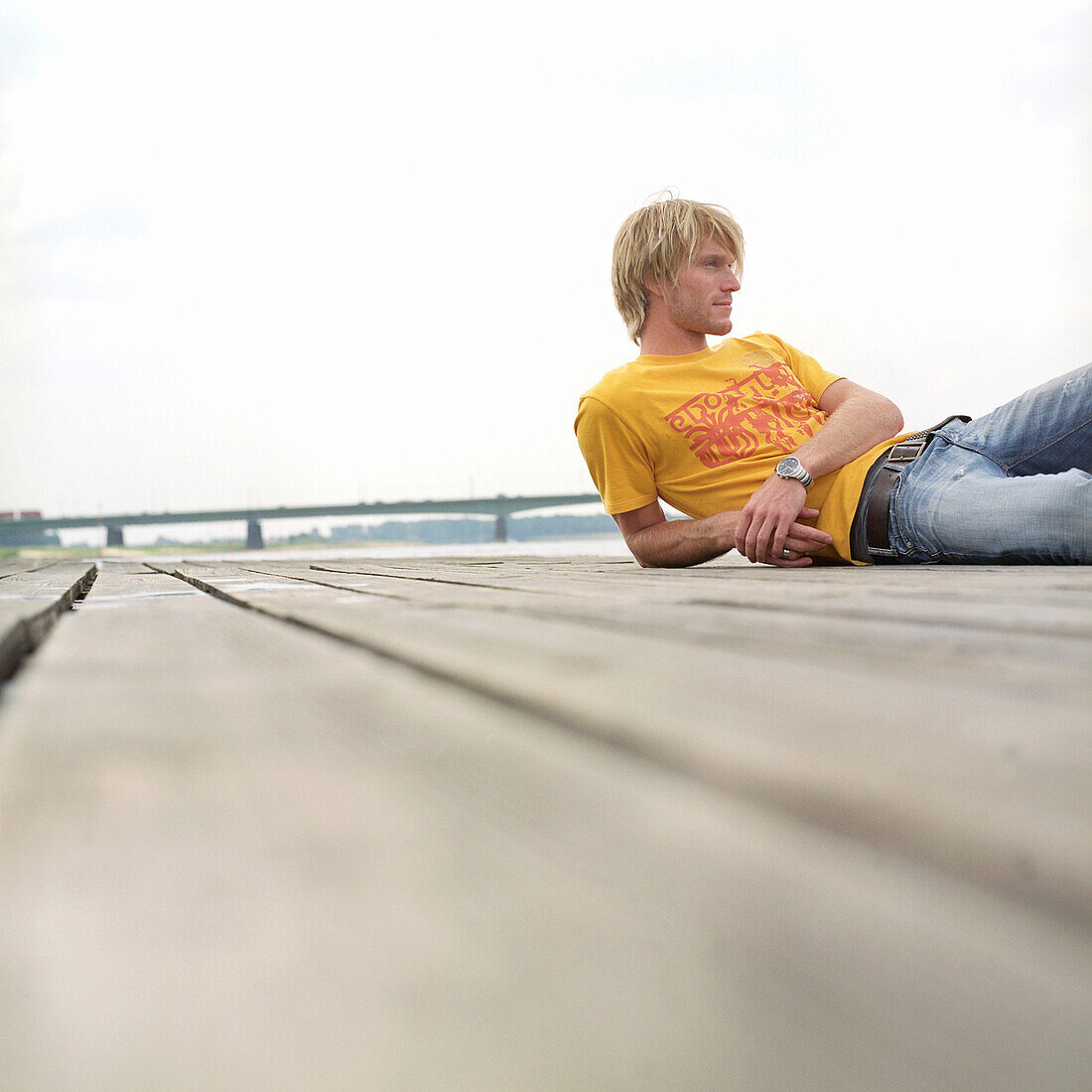 Mann liegt auf Holzboden, jung, blond, modisch, gelbes shirt, jeans, holzbolen, rhein, holzplattform, freizeit, entspannung, ruhe, deutscher