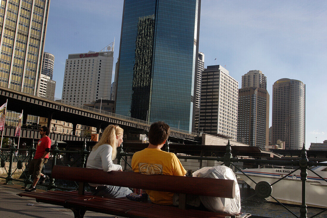 Touristen sitzen auf einer Bank auf den Kaianlagen, Circular Quay, Kai, Skyline des Geschäftsviertels, Central business district, CBD, Panorama, Hafen, Hauptstadt des Bundesstaates New South Wales, Sydney, Australien