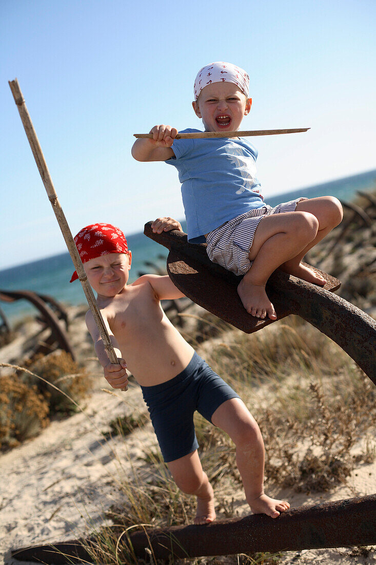 Zwei Jungen spielen Pirat auf einem rostigen Anker am Strand, Ilha de Tavira, Tavira, Algarve, Portugal