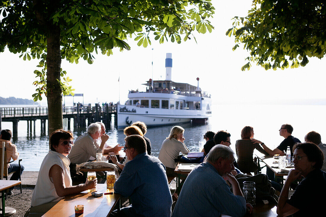 People sitting in a beer garden with steamboat and pier in the background, Herrsching, Ammersee, Bavaria, Germany